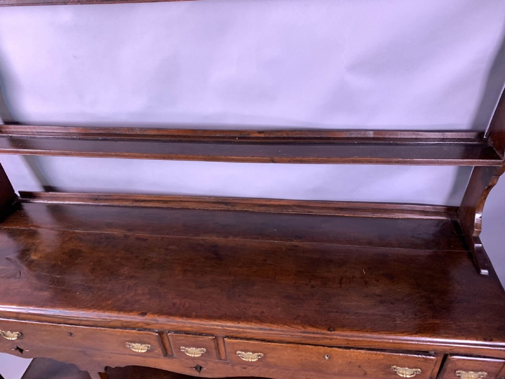 An 18th-century oak Welsh dresser featuring an open rack top with plate hooks, a central base with multiple drawers, and a lower pot board shelf. View XVIII