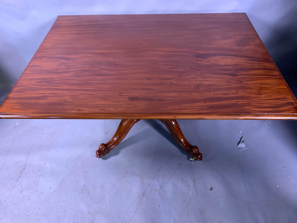 A rectangular mahogany breakfast dining table featuring a polished flame mahogany top set upon a central carved pedestal with splayed legs and brass castors.