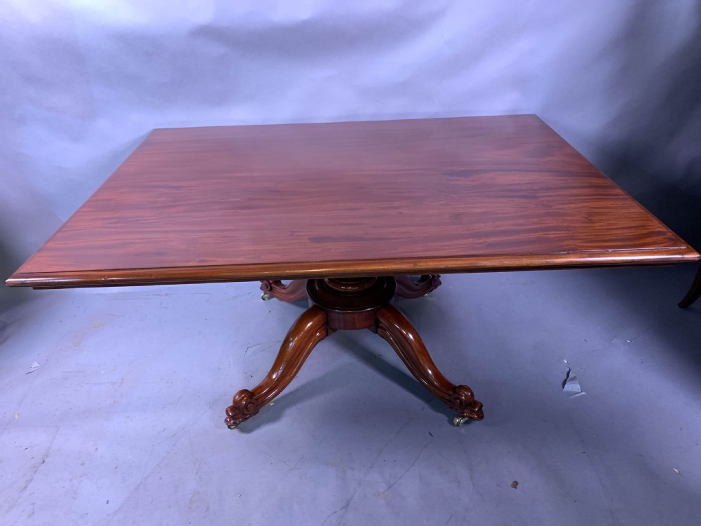 A rectangular mahogany breakfast dining table featuring a polished flame mahogany top set upon a central carved pedestal with splayed legs and brass castors. View II