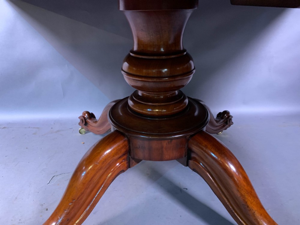 A rectangular mahogany breakfast dining table featuring a polished flame mahogany top set upon a central carved pedestal with splayed legs and brass castors. View IV