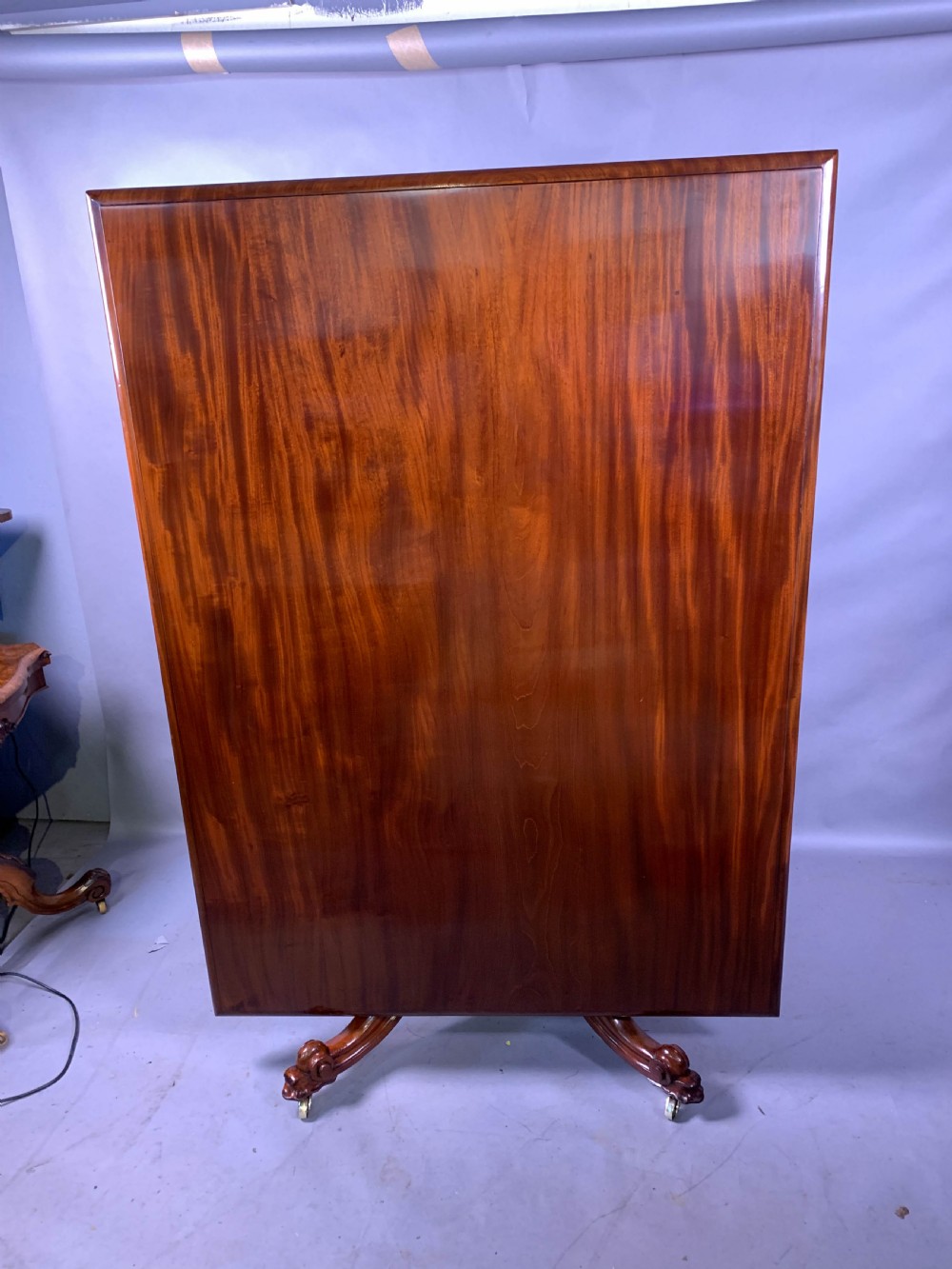 A rectangular mahogany breakfast dining table featuring a polished flame mahogany top set upon a central carved pedestal with splayed legs and brass castors. View XI