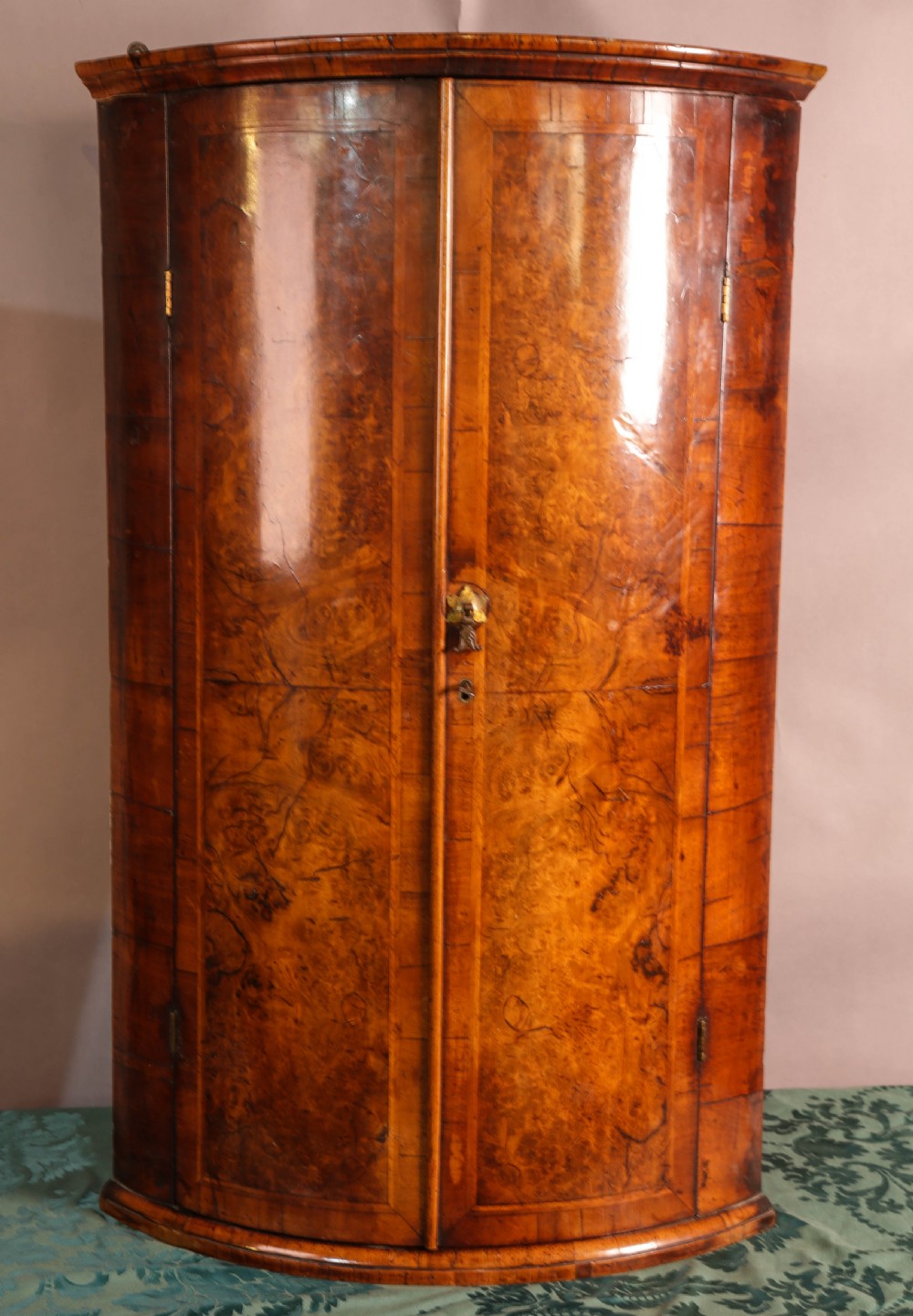 An eighteenth-century English bow-fronted corner cupboard crafted from figure-grained burr walnut with twin doors and a central brass escutcheon.