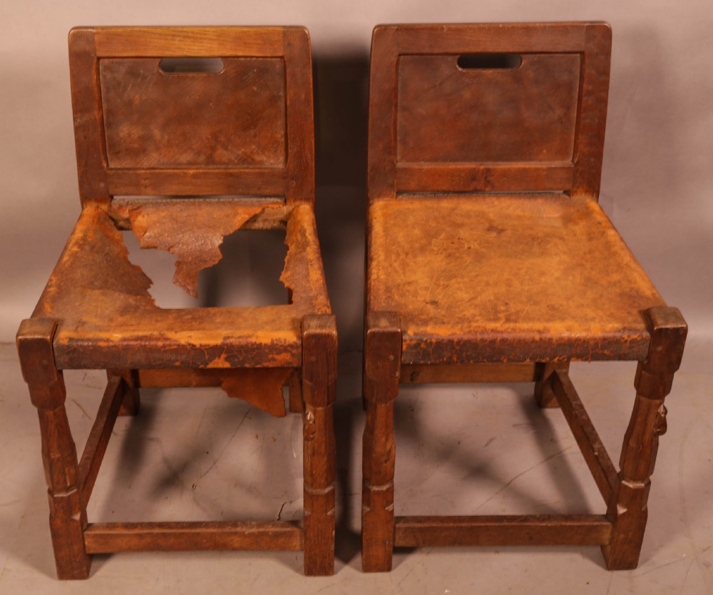 A pair of 1930s Arts and Crafts English oak side chairs featuring solid backs with cut-out handles, turned front legs with signature carved mouse motifs, and worn brown leather seats.
