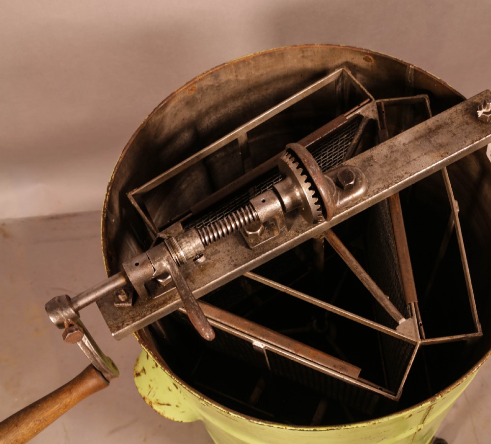 A pale green cylindrical metal honey separator with a top-mounted cast iron hand-crank mechanism and wooden handle. View V