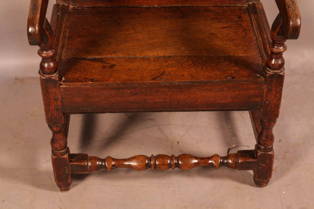 A 17th-century solid oak armchair featuring a panelled backrest with a shaped cresting rail, shaped arms, turned front legs, and plain stretchers. View V