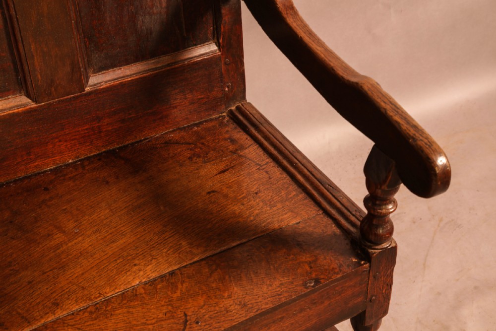 A 17th-century solid oak armchair featuring a panelled backrest with a shaped cresting rail, shaped arms, turned front legs, and plain stretchers. View VII