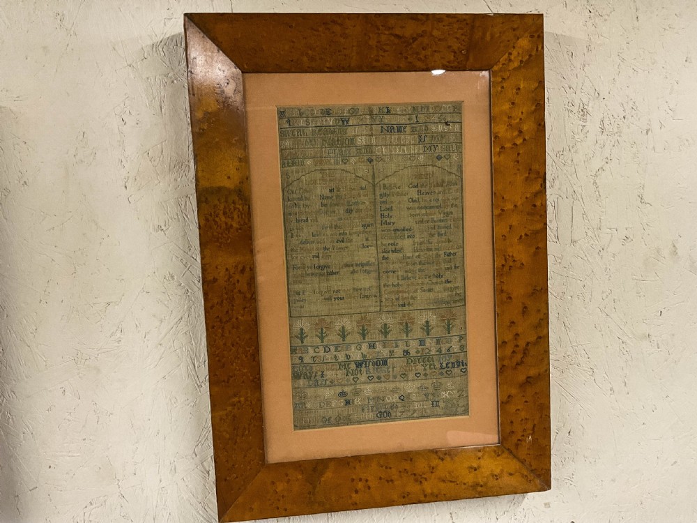An 18th-century framed needlework sampler featuring embroidered religious text, alphabets, and floral motifs within a wide burr wood frame. View II