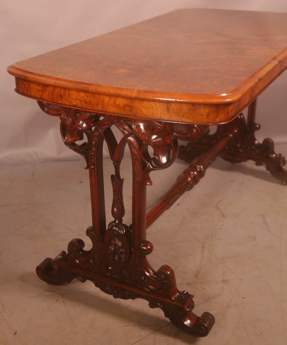 A Victorian burr walnut window table with a rectangular figured top, carved trestle supports, and a turned central stretcher. View VI