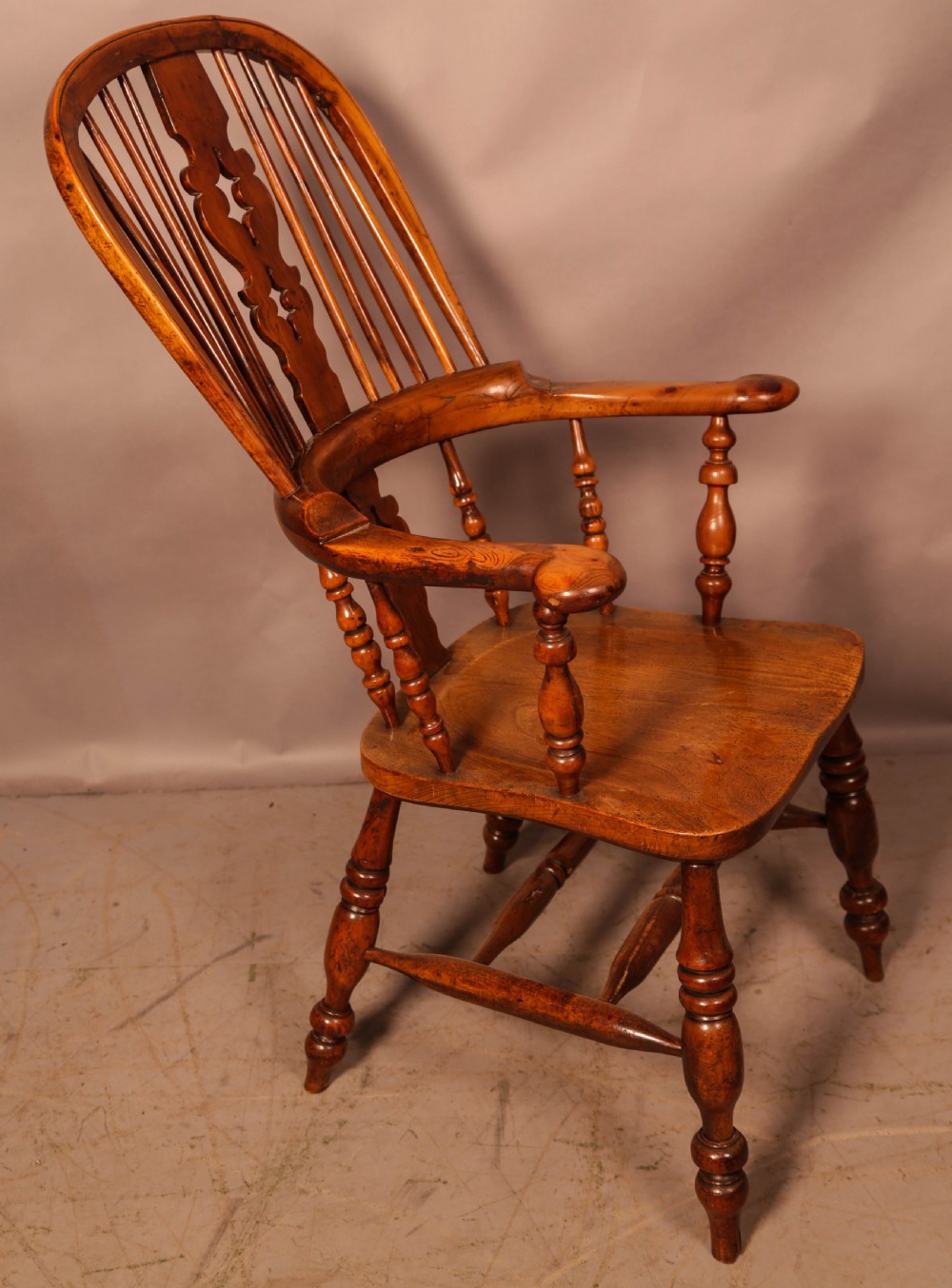 An antique English yew wood broad arm Windsor chair featuring a hooped back with a central pierced splat, turned legs, and a solid wood saddle seat.
