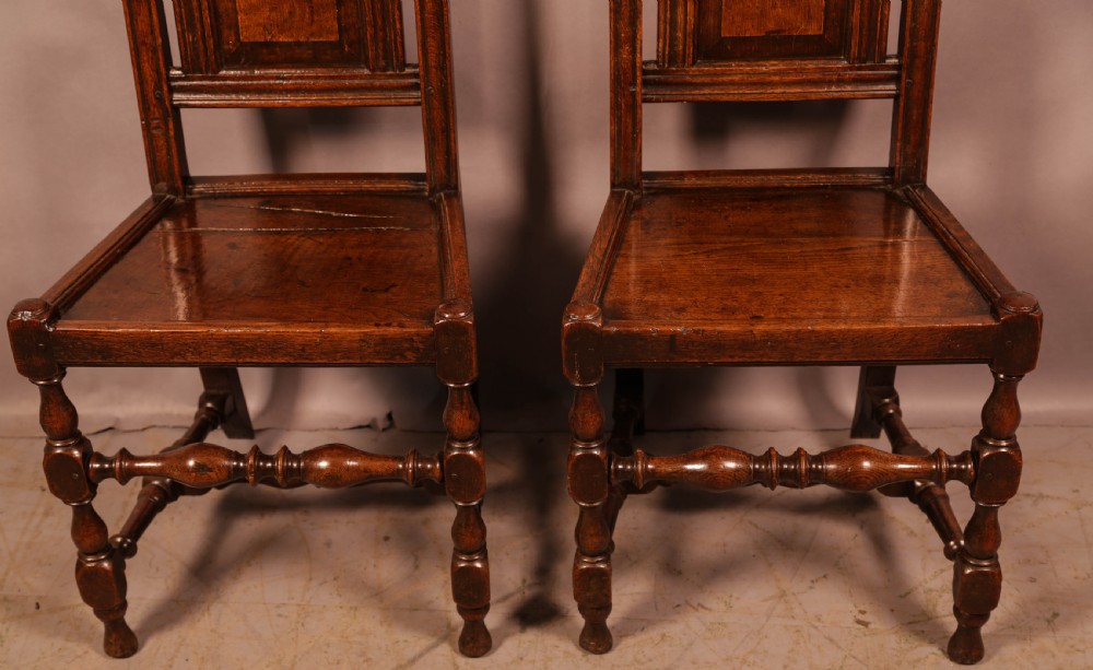 A pair of seventeenth-century oak hall chairs featuring high panelled backs with pierced scrollwork crests, solid timber seats, and turned front legs joined by stretchers. View II