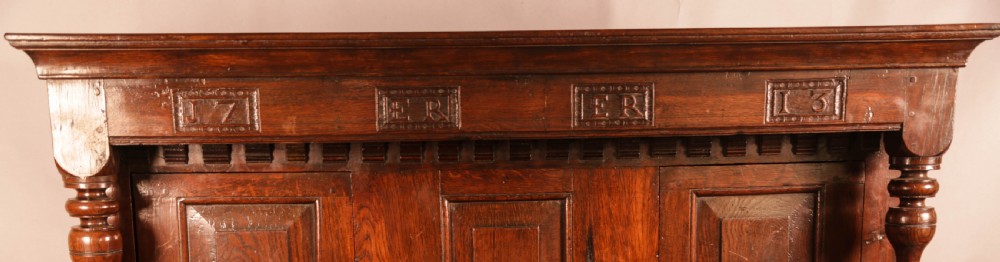 A front view of an 18th-century dark oak court cupboard featuring panelled doors, turned columns, and carved initials on the frieze. View IV