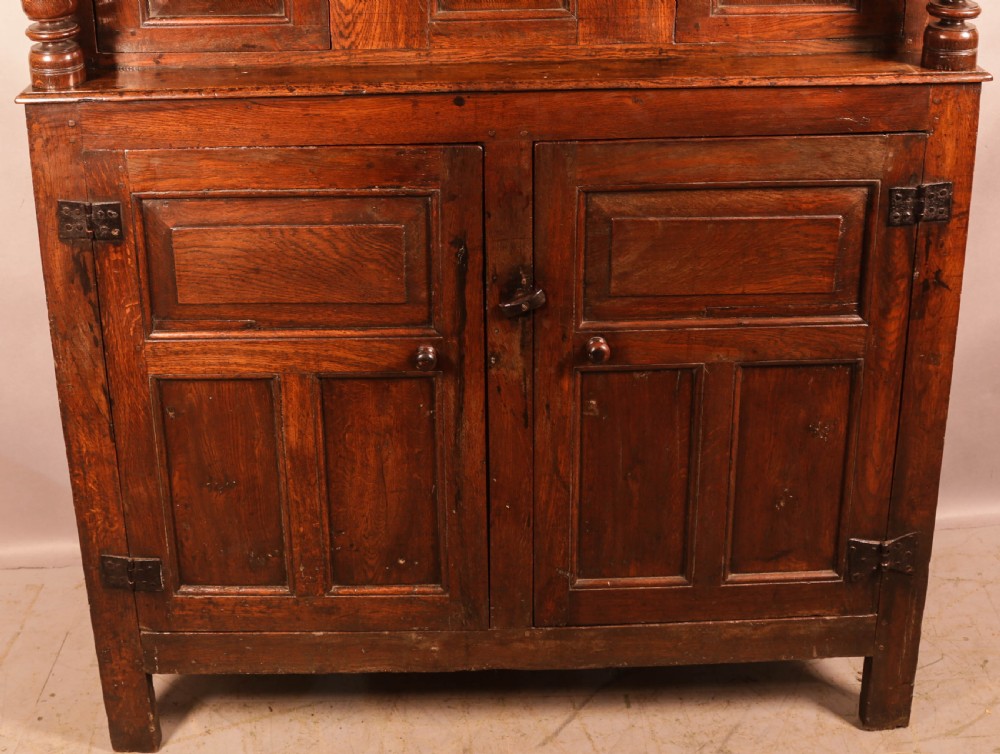 A front view of an 18th-century dark oak court cupboard featuring panelled doors, turned columns, and carved initials on the frieze. View V