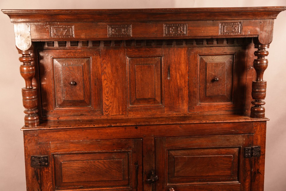 A front view of an 18th-century dark oak court cupboard featuring panelled doors, turned columns, and carved initials on the frieze. View VI