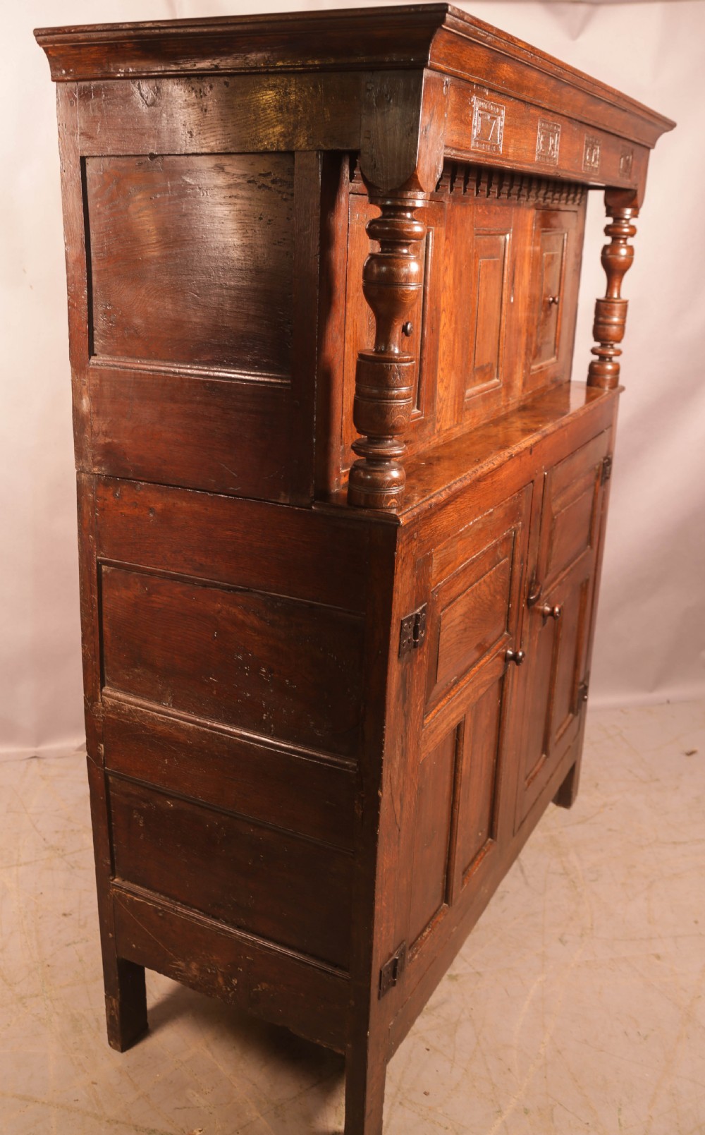 A front view of an 18th-century dark oak court cupboard featuring panelled doors, turned columns, and carved initials on the frieze. View XV