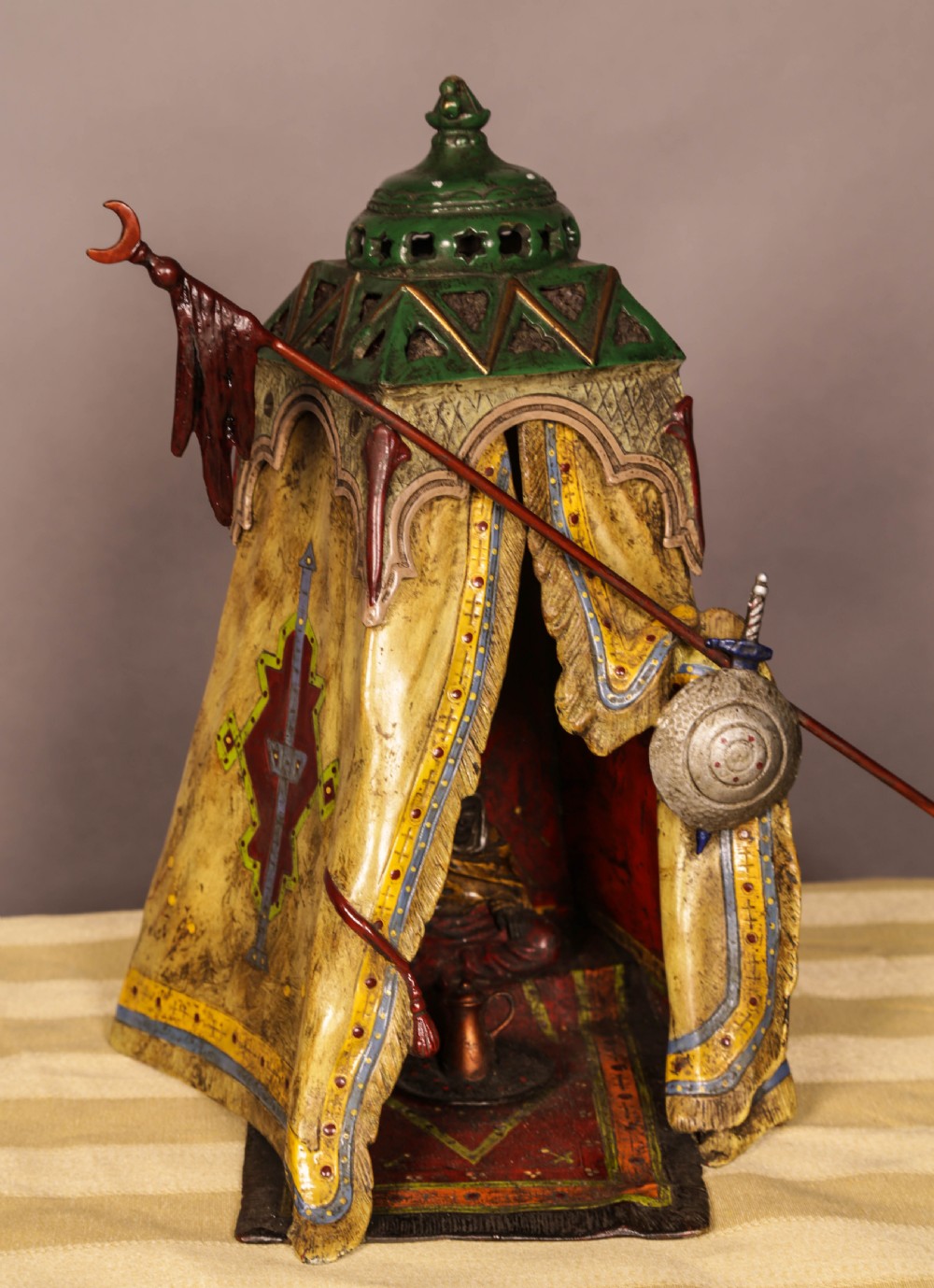 A cold-painted bronze sculpture depicting a bedouin tent with patterned yellow draping, a green domed top, and accompanying miniature weapons.