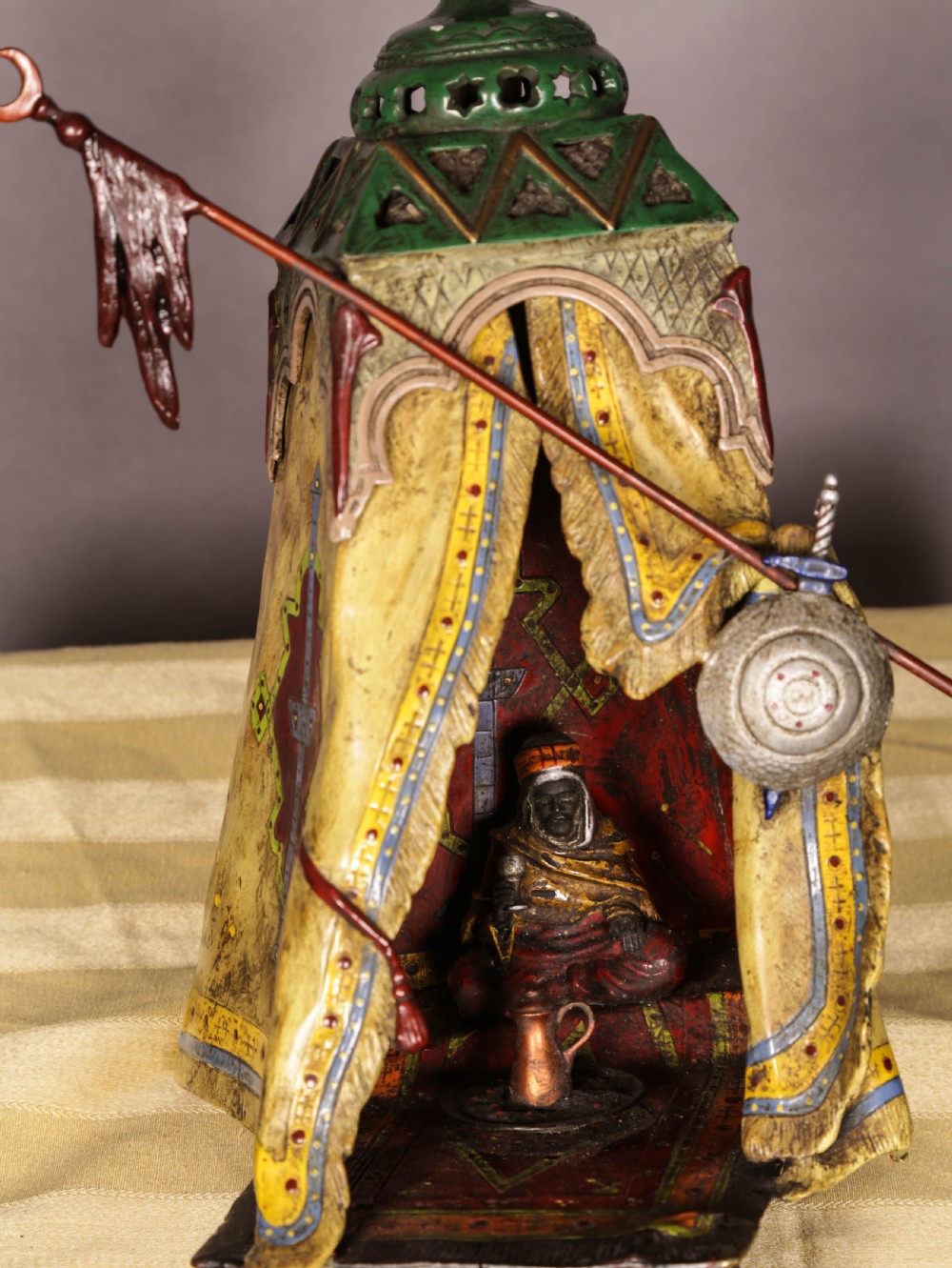 A cold-painted bronze sculpture depicting a bedouin tent with patterned yellow draping, a green domed top, and accompanying miniature weapons. View III
