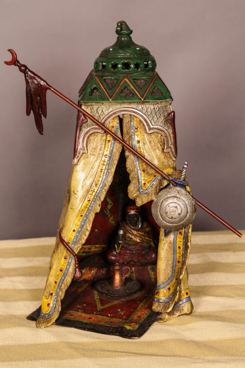 A cold-painted bronze sculpture depicting a bedouin tent with patterned yellow draping, a green domed top, and accompanying miniature weapons. View IX