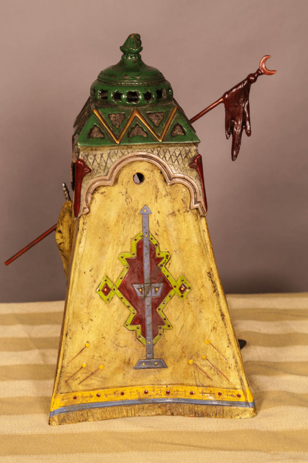 A cold-painted bronze sculpture depicting a bedouin tent with patterned yellow draping, a green domed top, and accompanying miniature weapons. View X