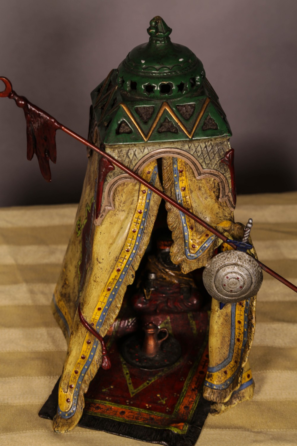 A cold-painted bronze sculpture depicting a bedouin tent with patterned yellow draping, a green domed top, and accompanying miniature weapons. View XIII