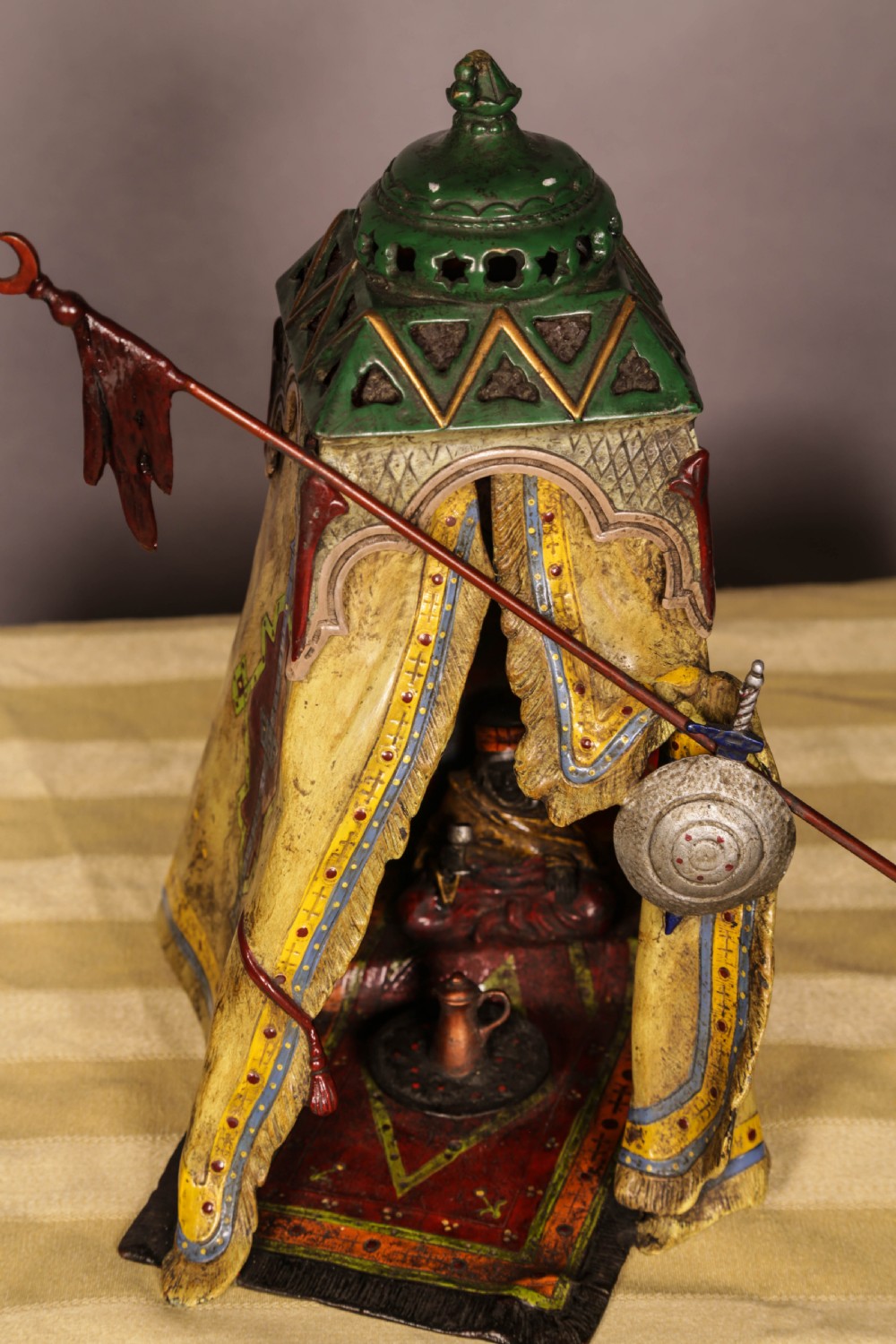 A cold-painted bronze sculpture depicting a bedouin tent with patterned yellow draping, a green domed top, and accompanying miniature weapons. View XIV