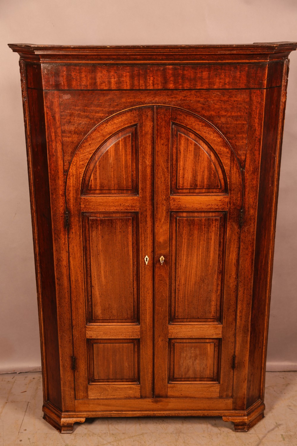 An 18th-century freestanding mahogany corner cupboard featuring arched double panel doors, keyhole escutcheons, and bracket feet.