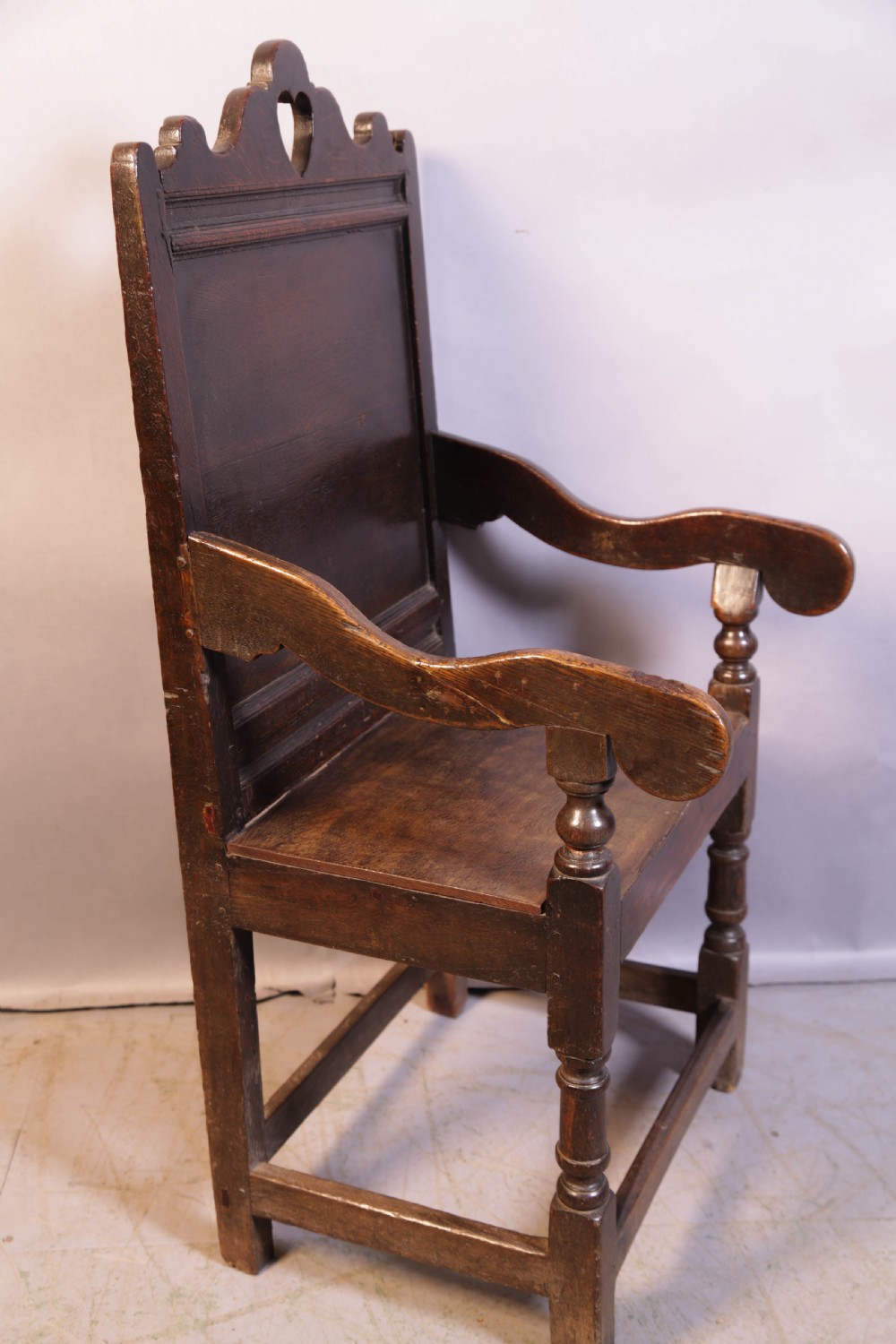 A dark oak 17th-century wainscot armchair featuring a panelled backrest with a shaped crest rail, curved armrests, and turned front legs.