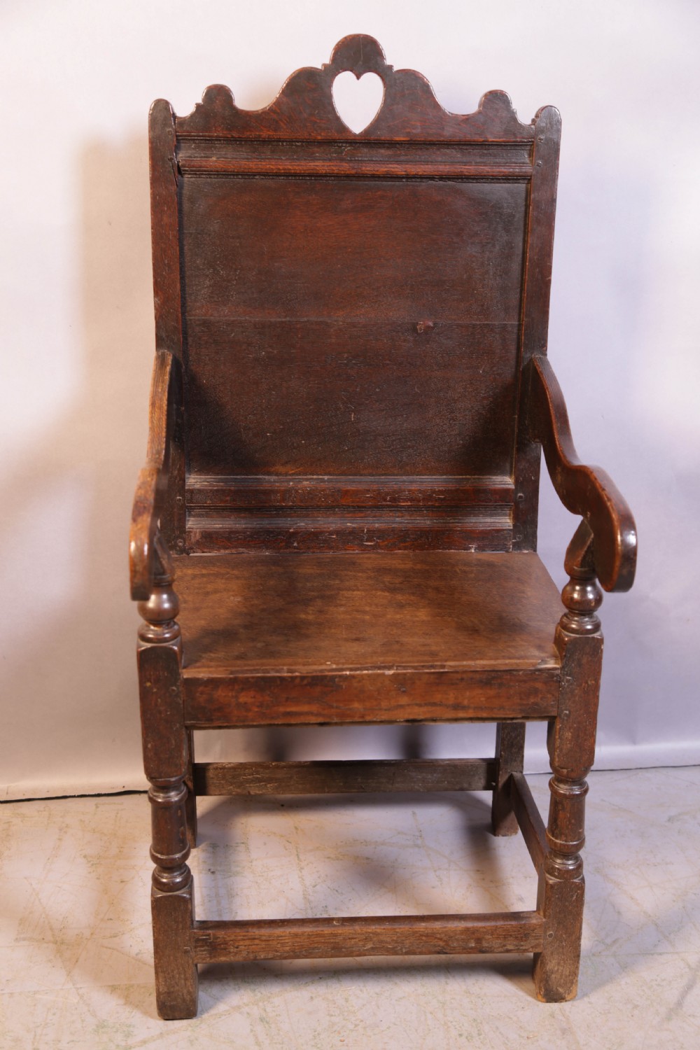 A dark oak 17th-century wainscot armchair featuring a panelled backrest with a shaped crest rail, curved armrests, and turned front legs. View II