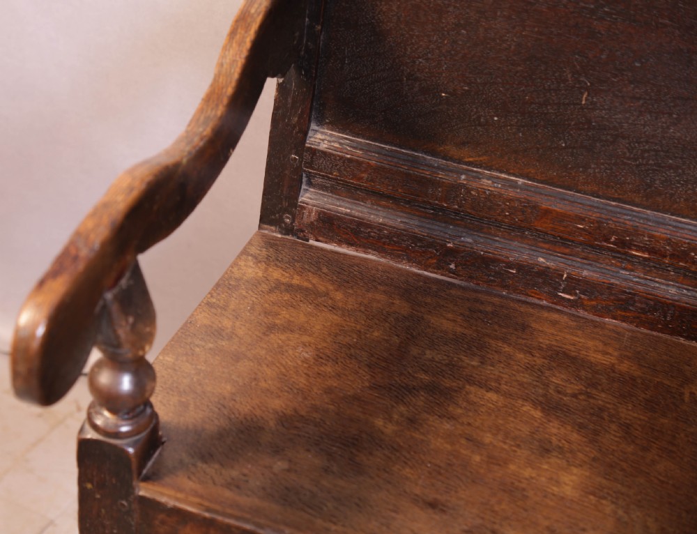 A dark oak 17th-century wainscot armchair featuring a panelled backrest with a shaped crest rail, curved armrests, and turned front legs. View V