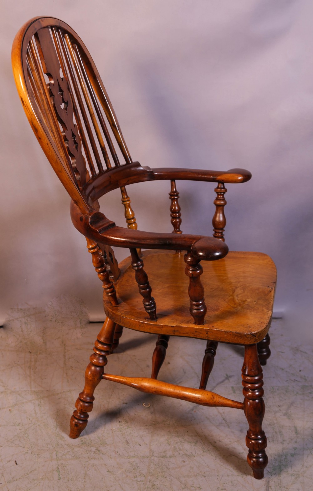 A warm brown yew wood Windsor chair with a hoop back, central pierced splat, wide armrests, turned legs, and H-stretcher supports.