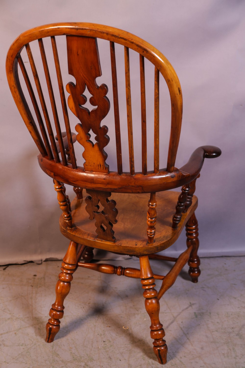 A warm brown yew wood Windsor chair with a hoop back, central pierced splat, wide armrests, turned legs, and H-stretcher supports. View II