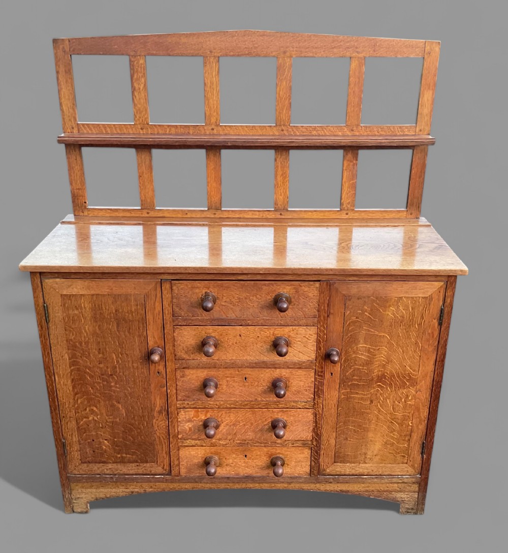 An Arts and Crafts solid oak dresser featuring a central column of five drawers flanked by panelled cupboard doors and an open grid-frame plate rack.