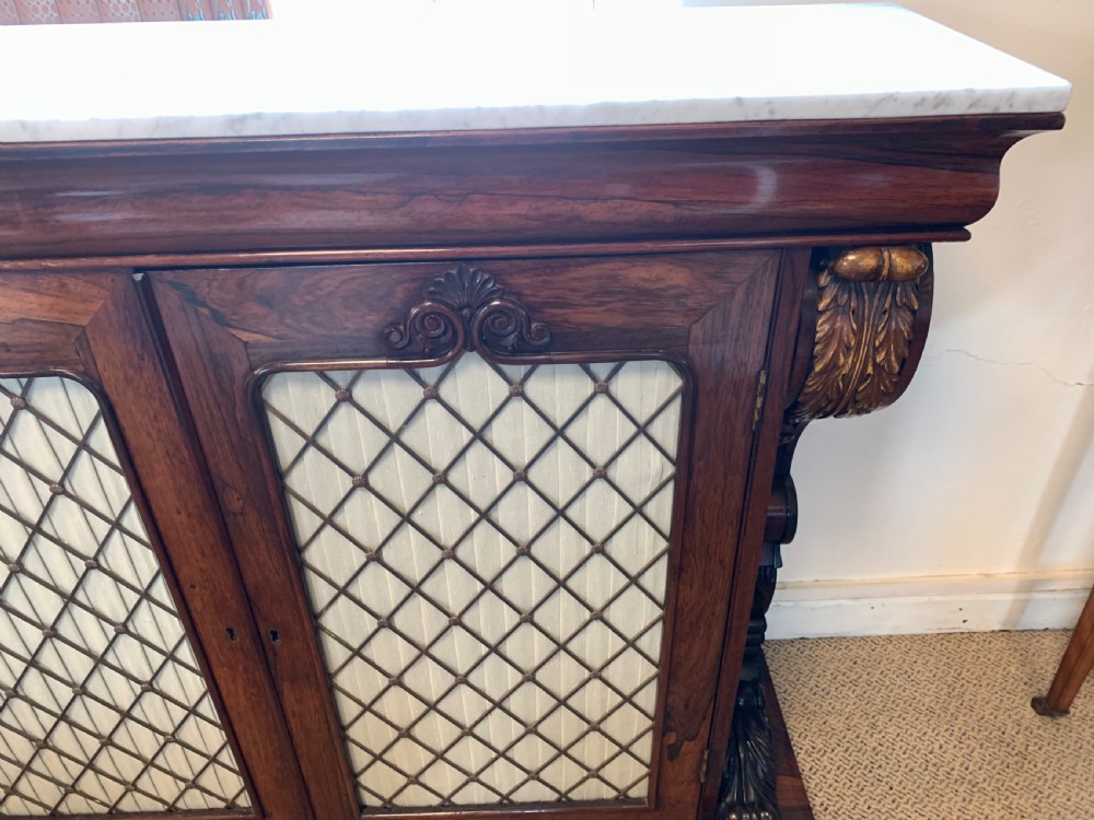 A dark rosewood chiffonier featuring a white marble top, carved gilt corbels, and two cupboard doors with pleated fabric behind a wooden lattice. View IX