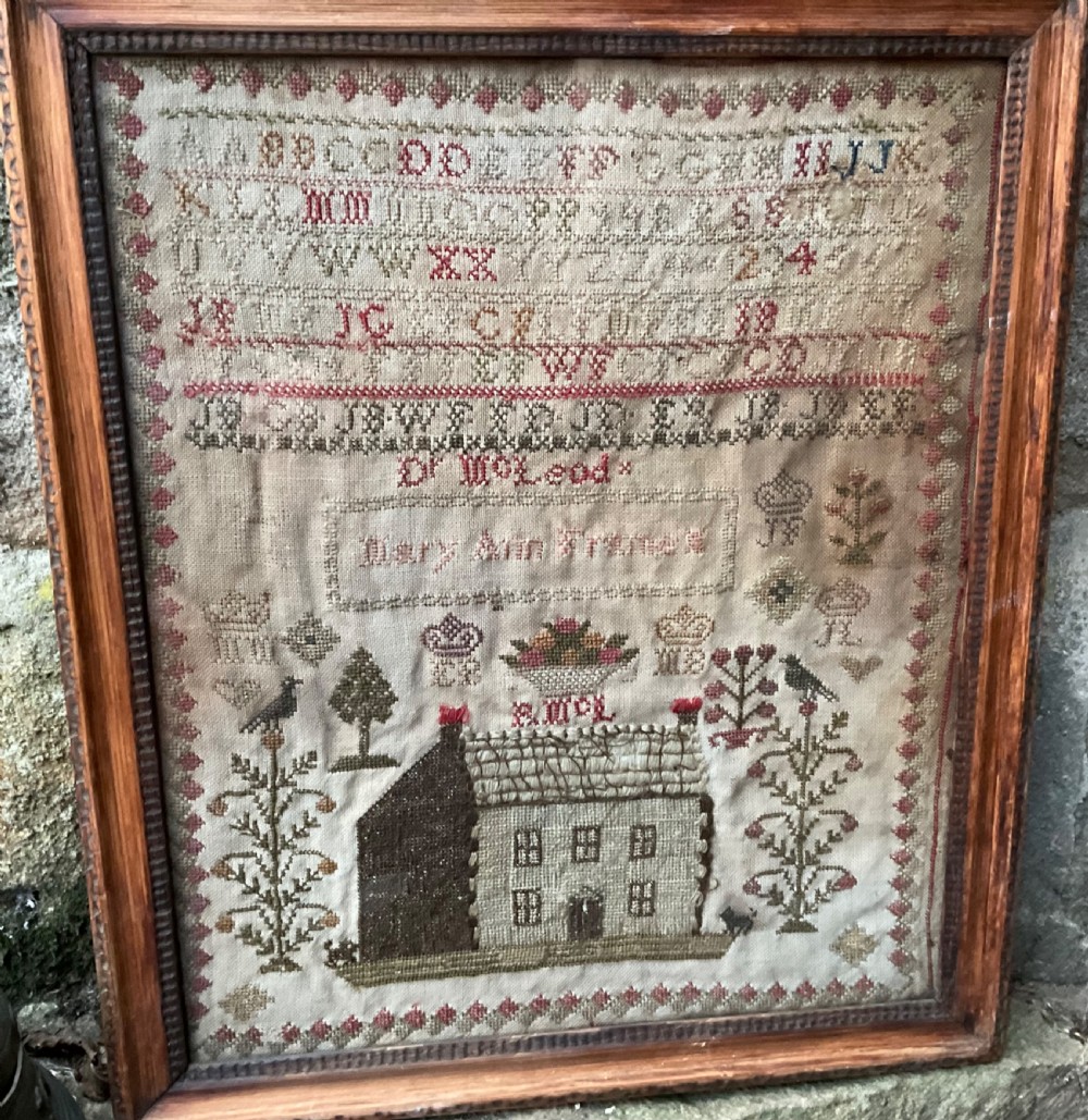 A framed antique needlework sampler on linen featuring stitched alphabets, numerals, architectural motifs, and a house, enclosed in a wooden frame.