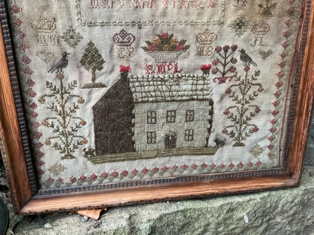 A framed antique needlework sampler on linen featuring stitched alphabets, numerals, architectural motifs, and a house, enclosed in a wooden frame. View II
