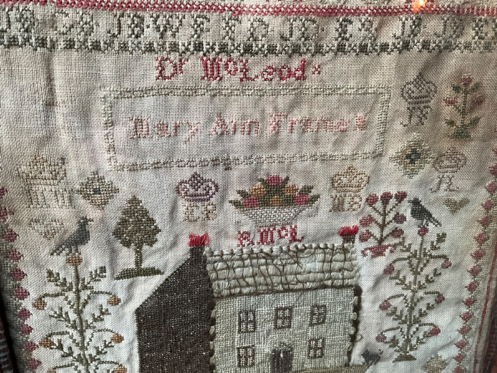 A framed antique needlework sampler on linen featuring stitched alphabets, numerals, architectural motifs, and a house, enclosed in a wooden frame. View IV