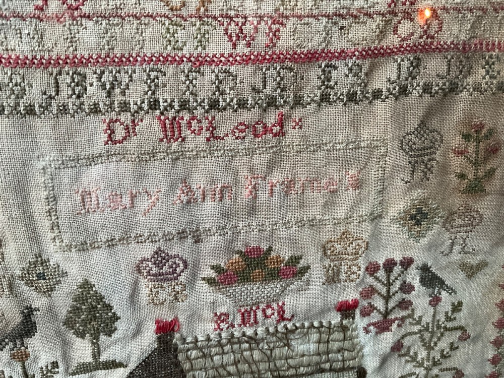 A framed antique needlework sampler on linen featuring stitched alphabets, numerals, architectural motifs, and a house, enclosed in a wooden frame. View V