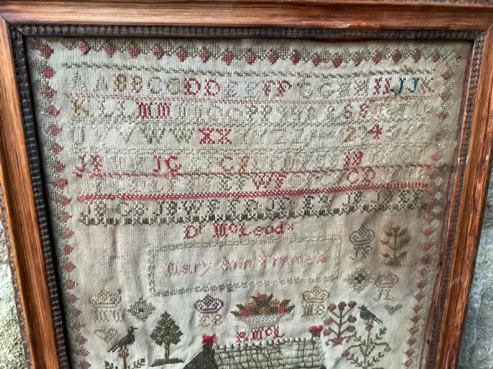 A framed antique needlework sampler on linen featuring stitched alphabets, numerals, architectural motifs, and a house, enclosed in a wooden frame. View VI
