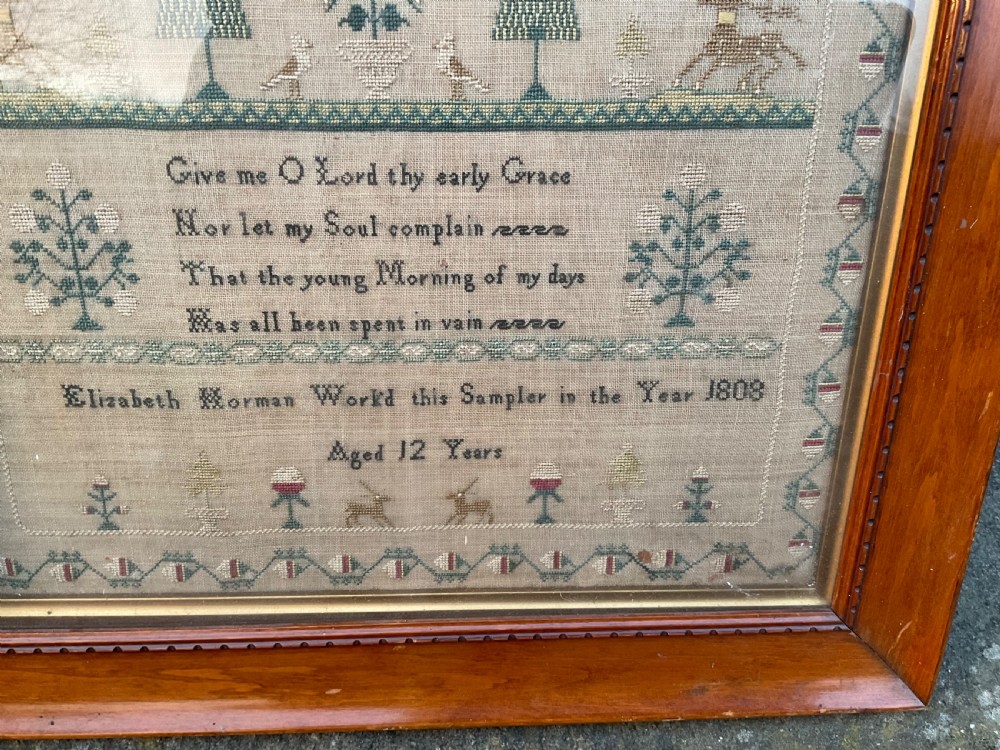 A framed antique linen needlework sampler featuring stitched text, geometric trees, animals, and decorative borders. View II