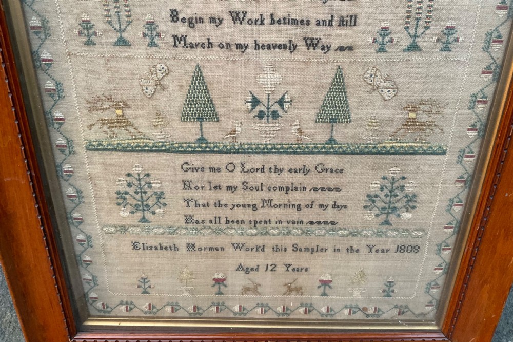 A framed antique linen needlework sampler featuring stitched text, geometric trees, animals, and decorative borders. View IV