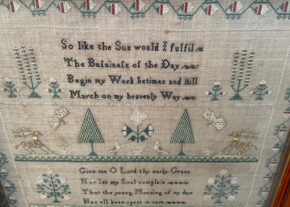A framed antique linen needlework sampler featuring stitched text, geometric trees, animals, and decorative borders. View VI