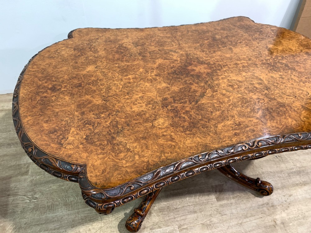 A rectangular Victorian burr walnut centre table with a figured top, carved edges, and a multi-scroll pedestal base. View V
