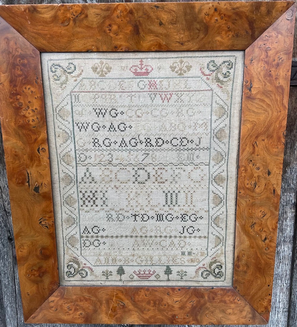 An antique hand-stitched needlework sampler displaying alphabets, numerals, coronet motifs, and the name Ann B Gillies inside a broad burr wood frame.