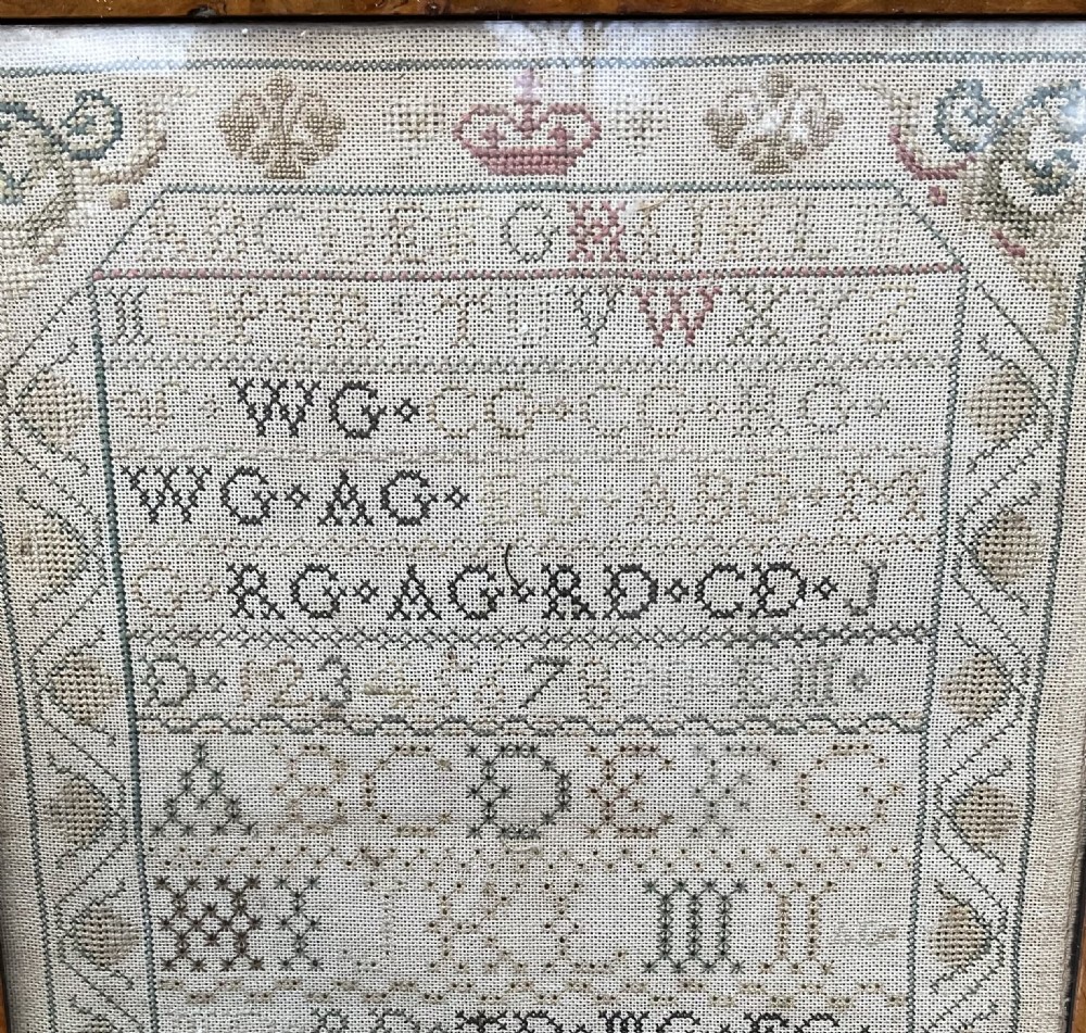 An antique hand-stitched needlework sampler displaying alphabets, numerals, coronet motifs, and the name Ann B Gillies inside a broad burr wood frame. View III