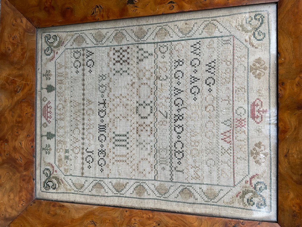 An antique hand-stitched needlework sampler displaying alphabets, numerals, coronet motifs, and the name Ann B Gillies inside a broad burr wood frame. View V