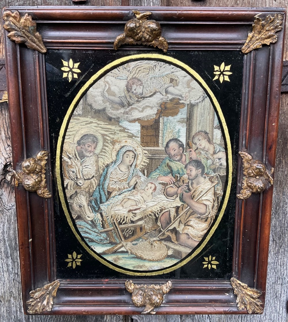 An eighteenth-century oval silk needlework depicting the Nativity scene, set within a black glass mat with gold star accents and a dark carved wood frame with gilt ornaments. View V
