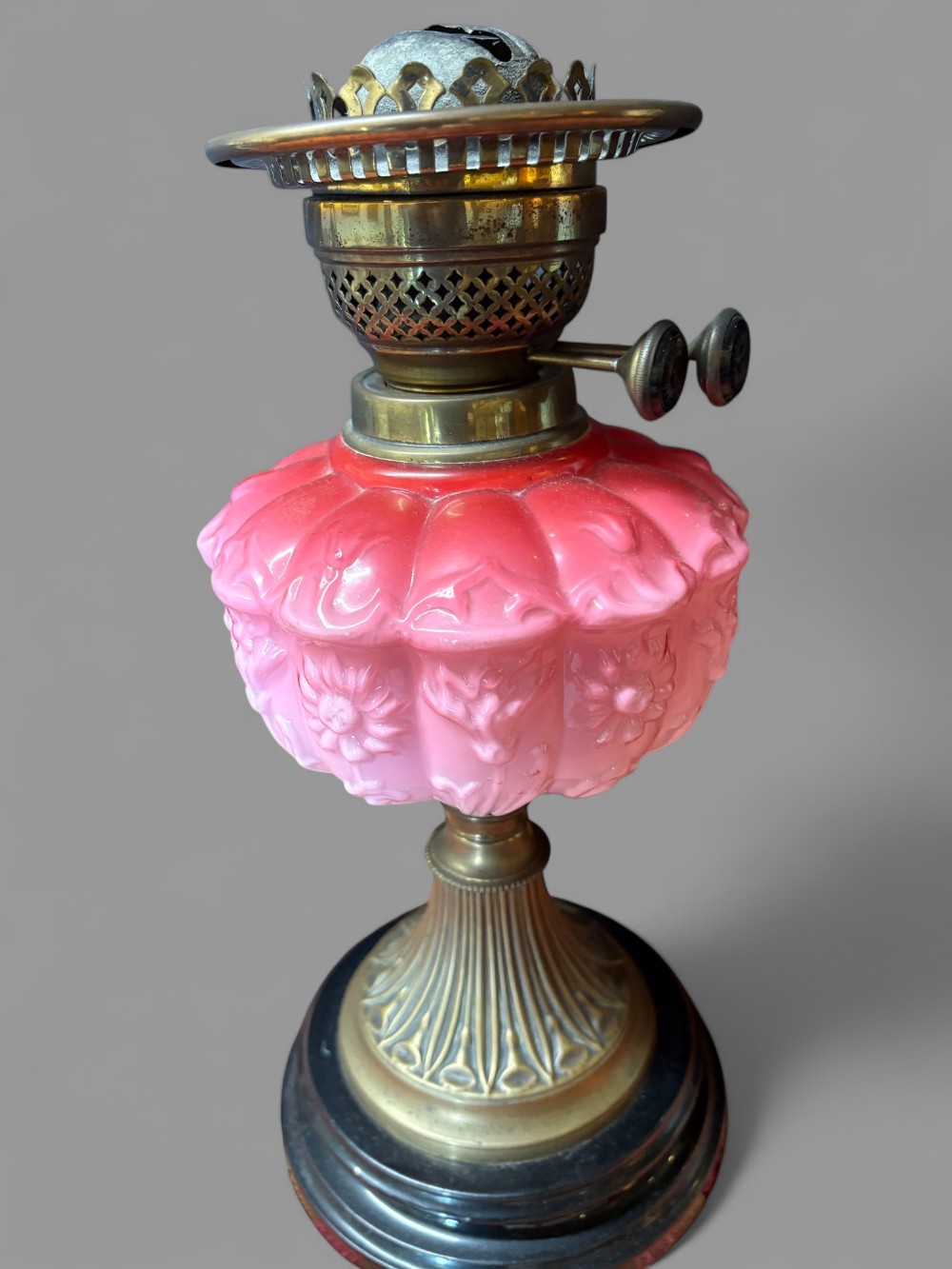 A Victorian oil lamp featuring a brass burner, a lobed pink opaque glass reservoir decorated with floral motifs, and a fluted brass column on a stepped black base.