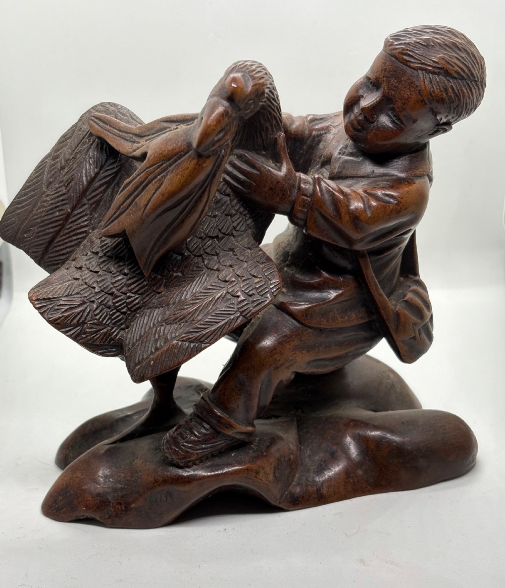 A hand-carved dark wood sculpture depicting a boy sitting alongside a large bird with detailed wing carving. View VII