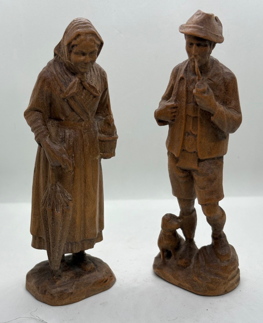 A pair of hand-carved wooden figurines depicting a man smoking a pipe with a small dog alongside a woman holding a basket and umbrella.