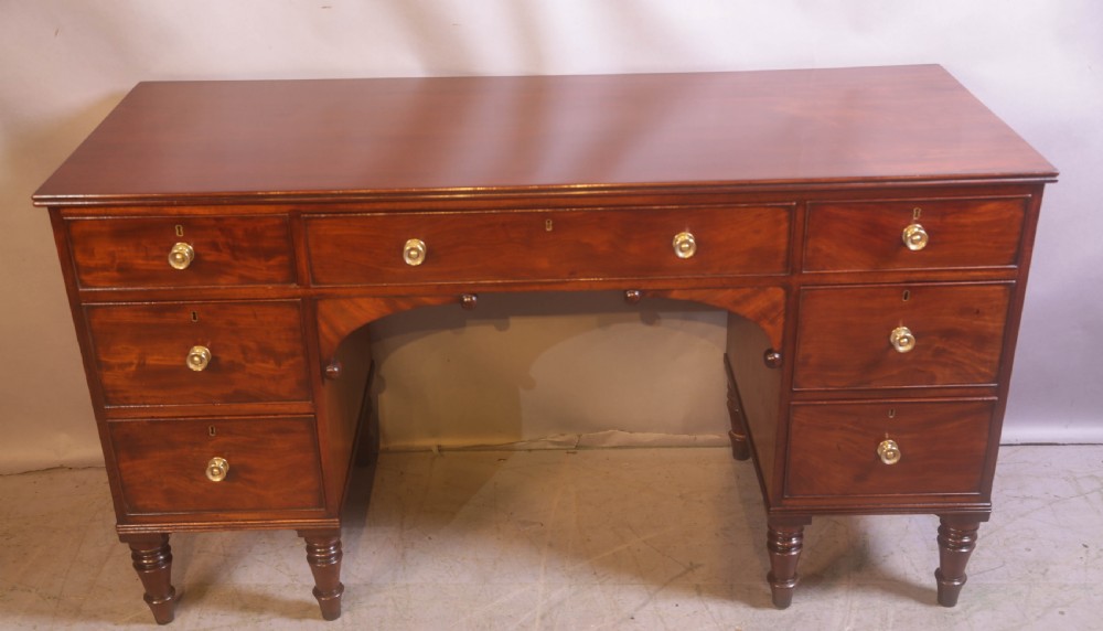 A 19th-century mahogany kneehole desk featuring multiple drawers with brass knobs, a central arched apron, and turned legs.