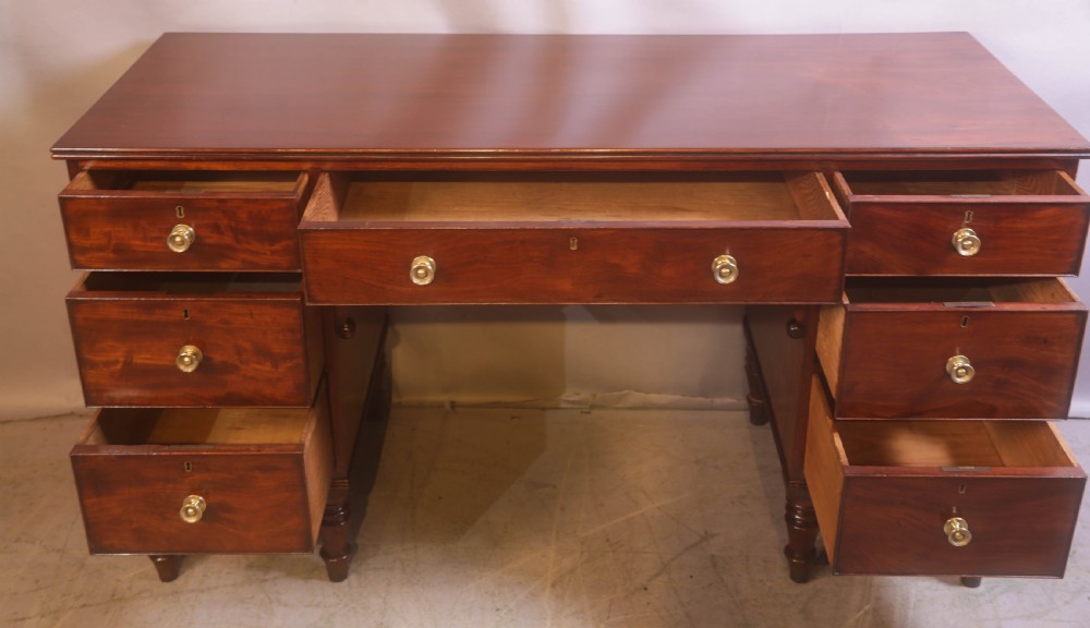 A 19th-century mahogany kneehole desk featuring multiple drawers with brass knobs, a central arched apron, and turned legs. View IV